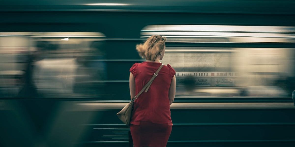 A woman in a red dress waits as a subway train speeds by in a blur, creating a sense of motion.