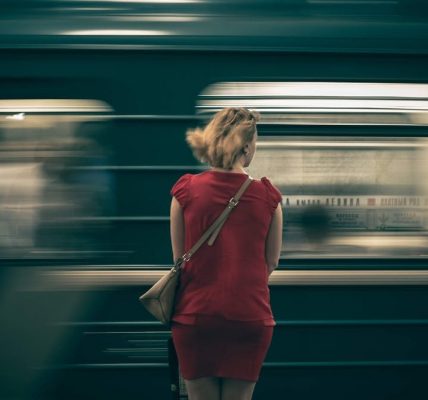 A woman in a red dress waits as a subway train speeds by in a blur, creating a sense of motion.
