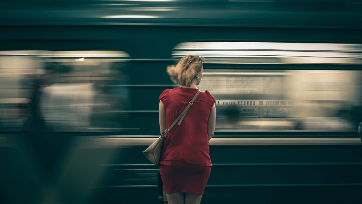 A woman in a red dress waits as a subway train speeds by in a blur, creating a sense of motion.